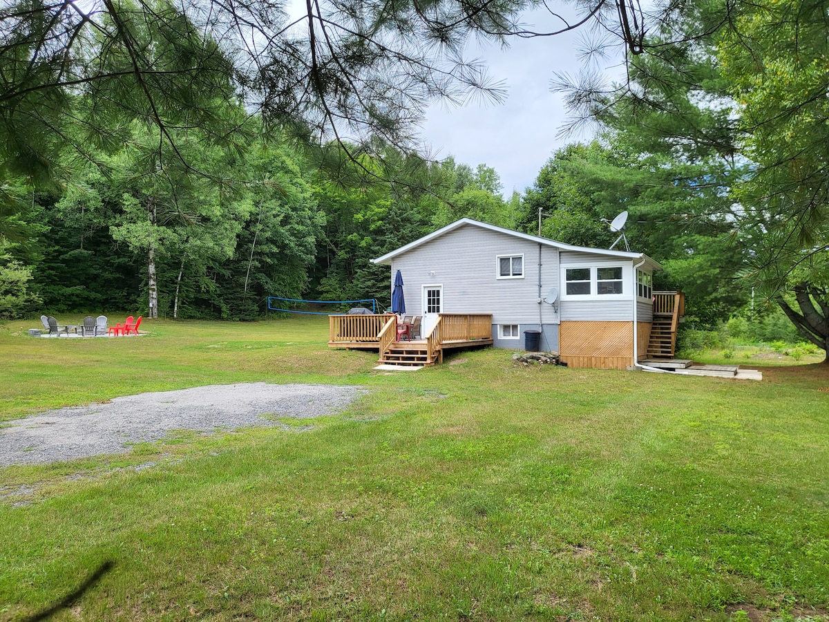 The cottage is situated on a spacious lawn surrounded by trees, with a wooden deck accessible by steps. A firepit area with seating is visible in the foreground, alongside a gravel path leading to the entrance. The peaceful setting emphasizes a connection to nature.