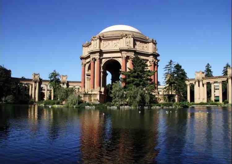 A grand architectural structure is reflected on a serene body of water, framed by lush greenery. The ornate building features a domed roof, tall arches, and intricate detailing, creating a harmonious blend with the surrounding nature and blue sky.