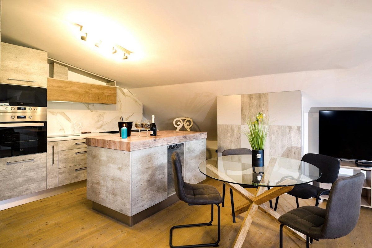 A modern kitchen area features sleek cabinetry and a central island with seating. A round dining table is accompanied by four stylish chairs, creating an inviting space for meals. Natural light enhances the warm wooden flooring and complements the contemporary decor.