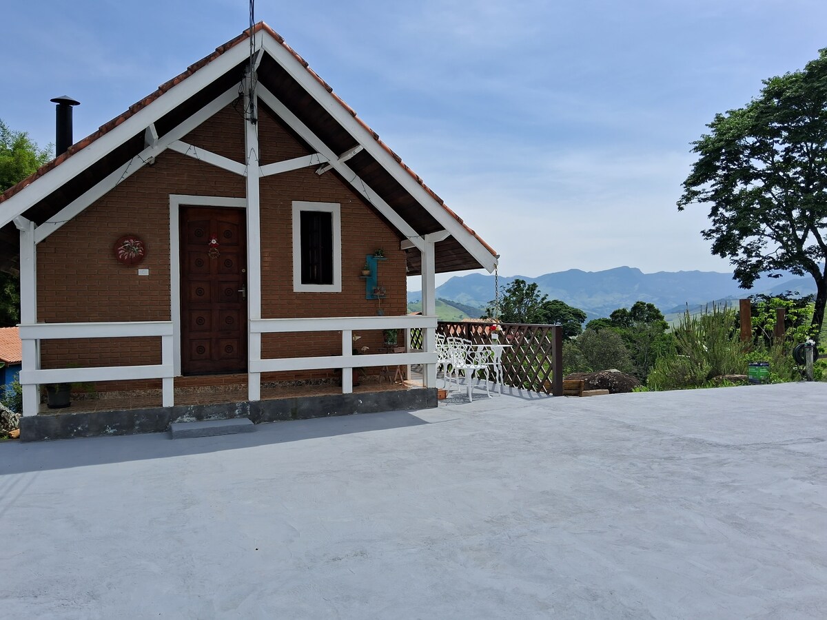The charming wooden chalet features a welcoming facade with a brown exterior and a sloped roof. Surrounding the chalet are lush green landscapes and mountain views in the background, offering a sense of tranquility. A small outdoor seating area is visible, providing space for relaxation.