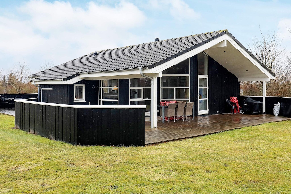 An exterior view of the holiday home displays a modern black structure with a distinctive angled roof. A spacious wooden terrace, furnished with a dining table and chairs, surrounds the house. Lush green grass and a subtle landscape provide a serene backdrop.