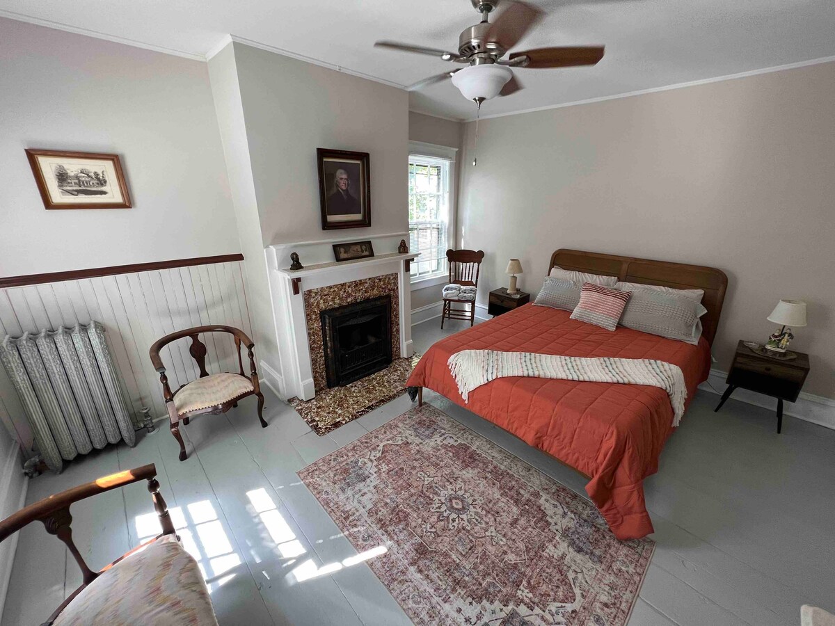 A cozy bedroom features a spacious bed with an orange-colored blanket, complemented by antique-style chairs and nightstands. A vintage fireplace adds character to the room, with sunlight illuminating the wood floor and enhancing the serene atmosphere.