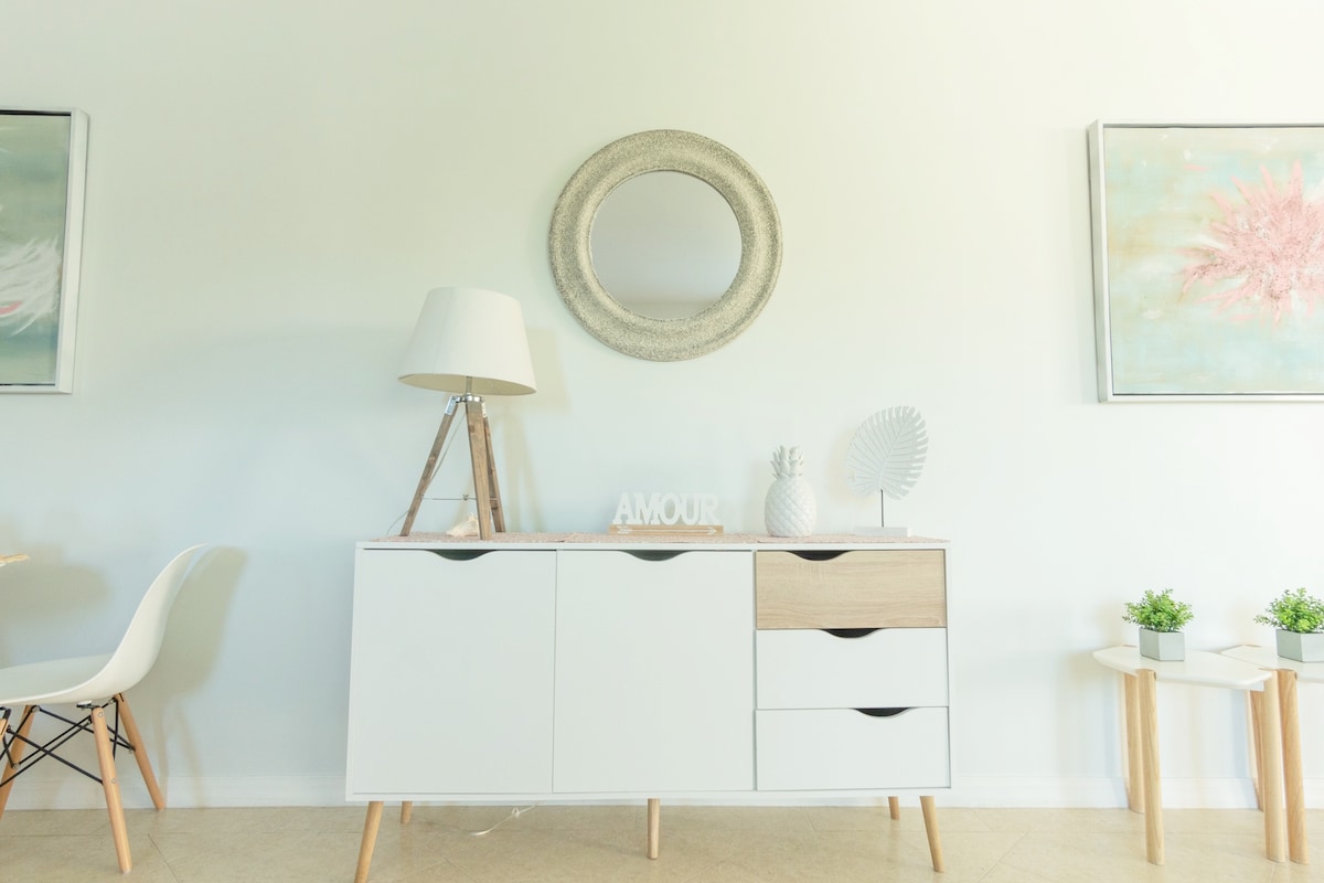 A modern sideboard in white and wood finishes is positioned against a light wall. A round mirror hangs above, reflecting soft light. The space is accented by two framed art pieces with pastel colors, a stylish lamp, and small potted plants placed on wooden stands.