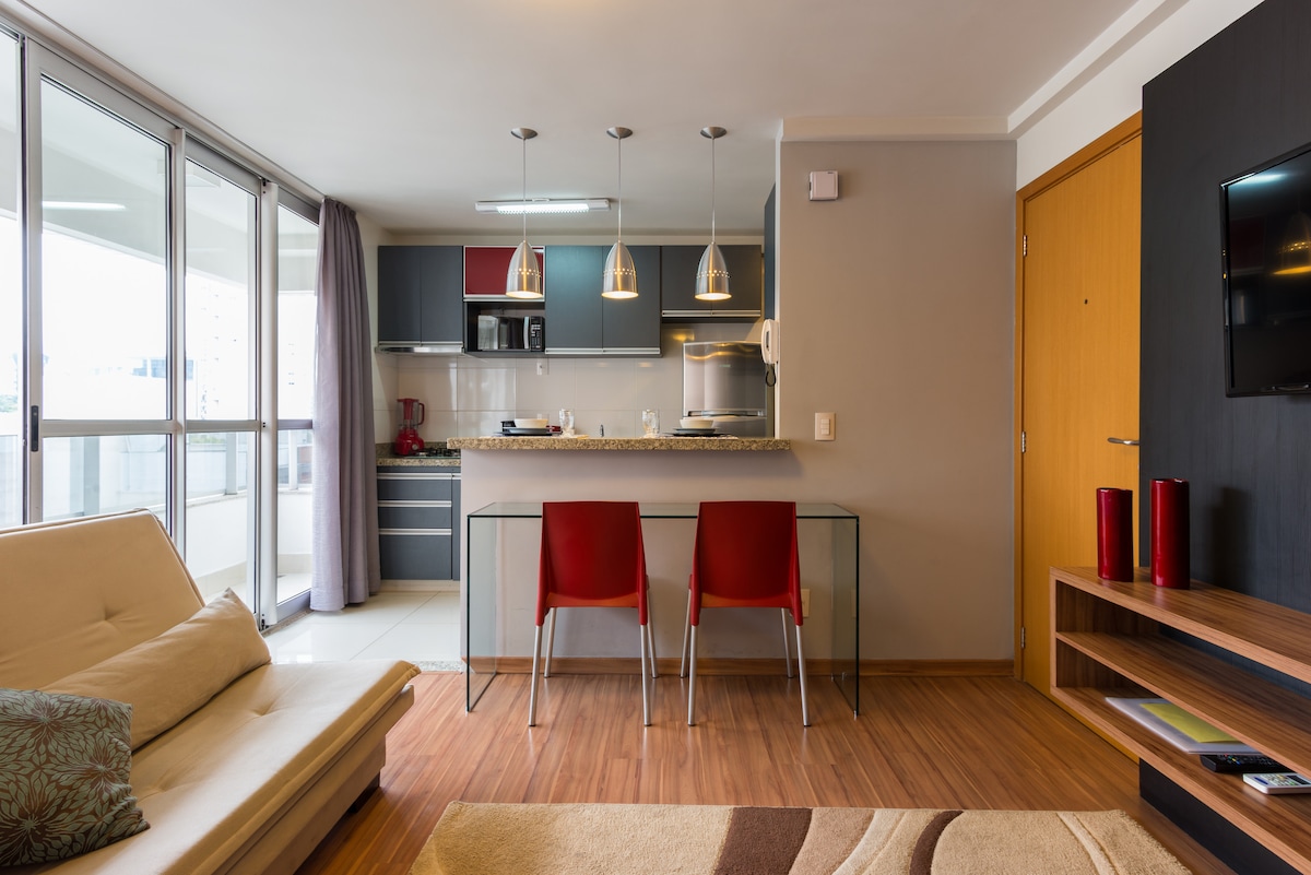 The open living space features a sleek, modern kitchen with stainless steel appliances and a granite countertop. A glass dining table with two red chairs is situated in front of a large window, allowing ample natural light to fill the area. A cozy sofa adds comfort to the room.