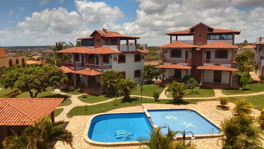 A landscaped area is depicted with several buildings featuring red-tiled roofs. A swimming pool, with a light blue hue, is centrally situated among palm trees and manicured grass. Walkways made of stone provide access to the various residences. The setting is open with a few clouds in the sky.