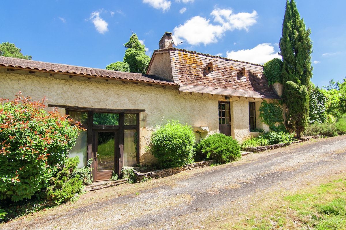 The exterior of the mansion features a rustic stone facade surrounded by lush greenery. A gravel pathway leads up to large doors framed by vibrant foliage, providing a welcoming entrance to the property.