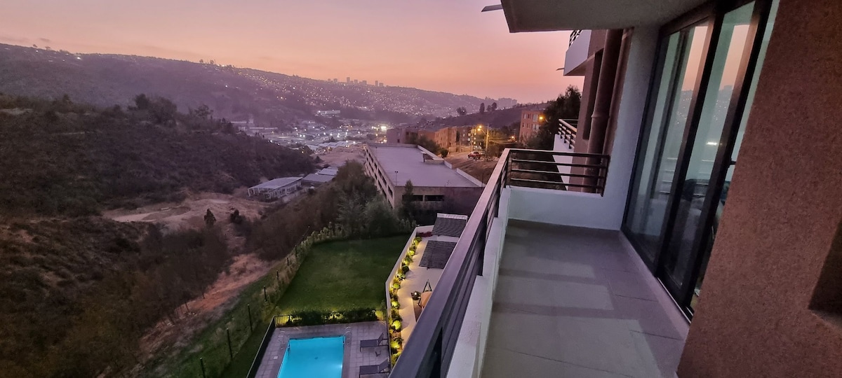 A spacious balcony is shown, offering a view of the swimming pool below and the surrounding hills at dusk. Soft outdoor lighting highlights the greenery along the edge of the property, while the distant city skyline is visible under a colorful evening sky.