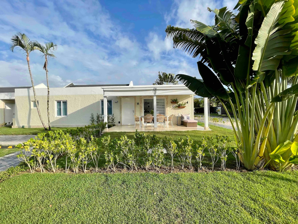 The exterior of a stylish home is framed by lush greenery and palm trees. A covered porch, furnished with delicate seating, invites relaxation. A well-maintained lawn contrasts with the light-colored facade, while a pathway leads to the entrance, enhancing the appeal of this tranquil setting.