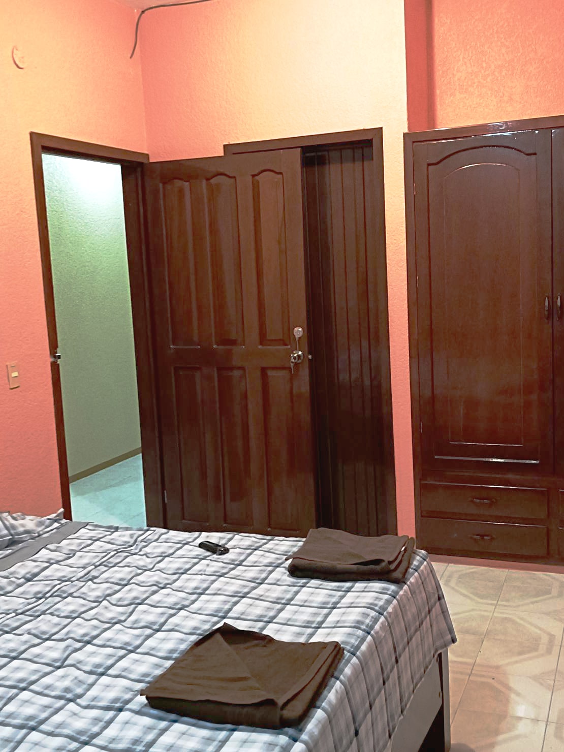 A bedroom features a spacious bed with a checkered blanket. Dark wooden doors are visible, leading to an adjoining room and a closet. Shelves and drawers are integrated into the closet unit, enhancing storage options. Soft orange walls create a warm ambiance.