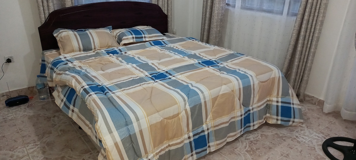 A spacious bed is layered with a checkered blanket in neutral tones, complemented by two matching pillows. Soft light filters through sheer curtains, illuminating the room. A clean floor and minimal furnishings create an uncluttered atmosphere.