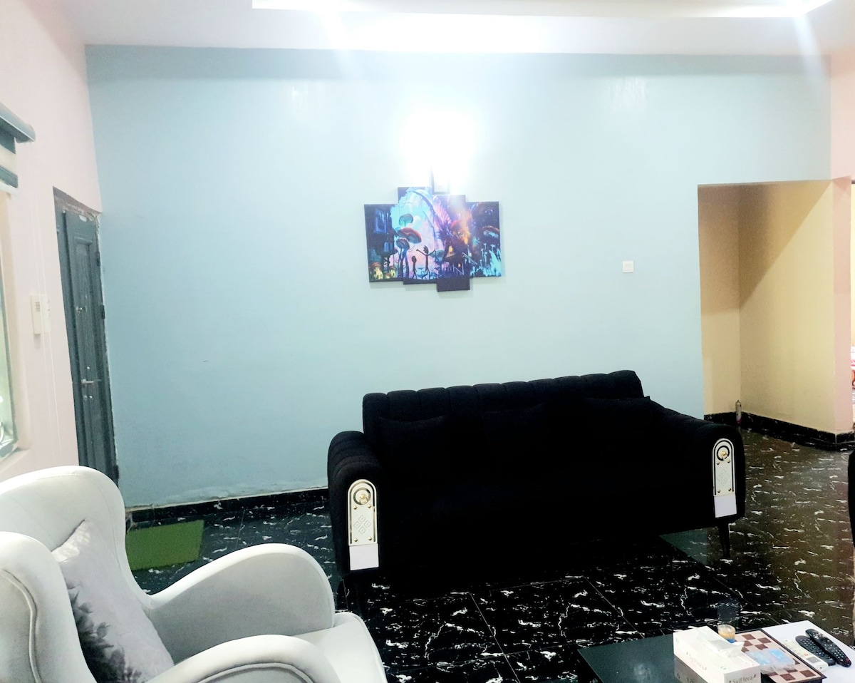 A living area with a black sofa and a light-colored armchair is presented. A television is mounted on the wall, displaying colorful artwork. The floor features dark, glossy tiles, and the walls are painted in a soothing blue hue.