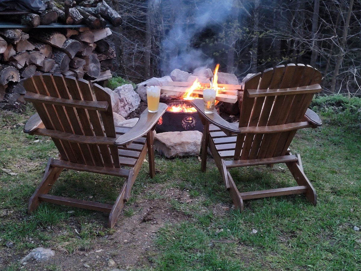 Two wooden Adirondack chairs are positioned facing a circular stone fire pit, where flames are seen glowing amidst glowing embers. Two glasses filled with a light beverage rest on the ground nearby. A stack of firewood is arranged in the background, surrounded by greenery.