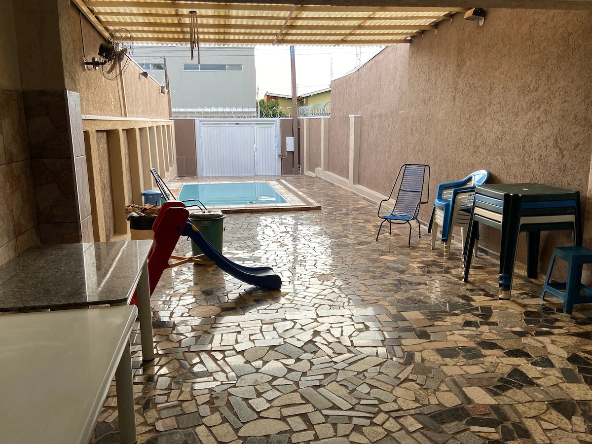 An outdoor area features a patterned stone floor, with a swimming pool partially visible at the far end. Seating options include two blue chairs and a green table, alongside a red and blue slide. Natural light brightens the space beneath a slatted roof.
