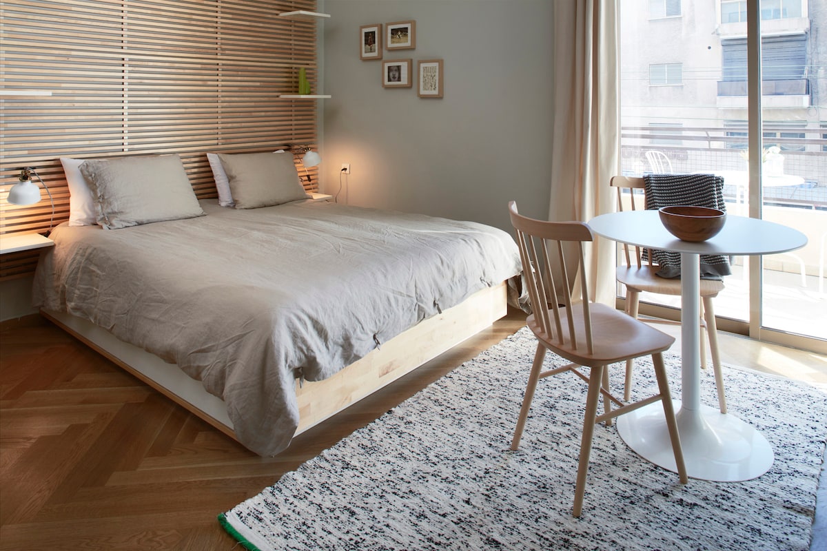 A bright bedroom features a spacious bed with light bedding, complemented by wooden accents. A small dining table with a chair is present, accompanied by a textured rug. Soft natural light streams through a sliding glass door, leading to an outdoor space.