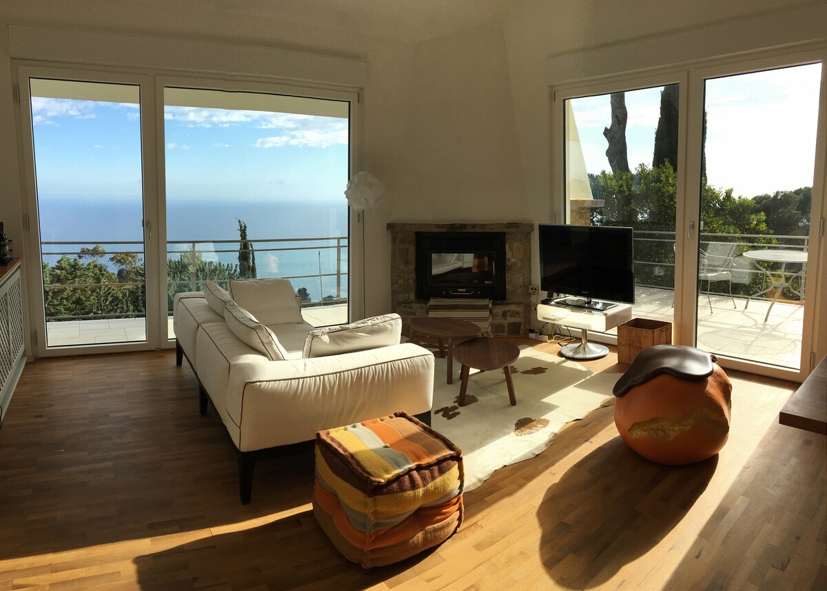 A bright living area is presented with large glass doors that open to a breathtaking sea view. A comfortable sofa is positioned near a stone fireplace, accompanied by a television and a wooden coffee table. Colorful poufs add a touch of warmth to the space.