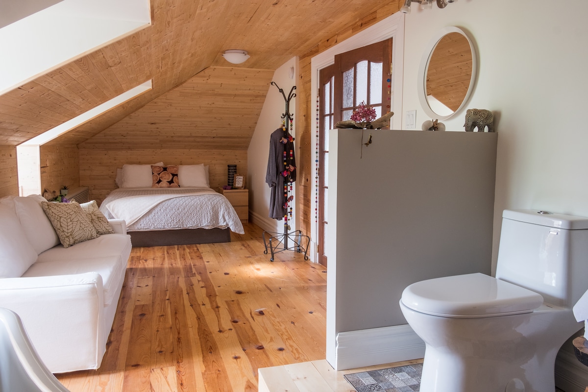 The room features sloped wooden ceilings and natural light streaming through a window. A spacious bed with neutral bedding is positioned near a plush sofa. A partitioned area contains a modern toilet, with wooden flooring enhancing the cozy atmosphere.