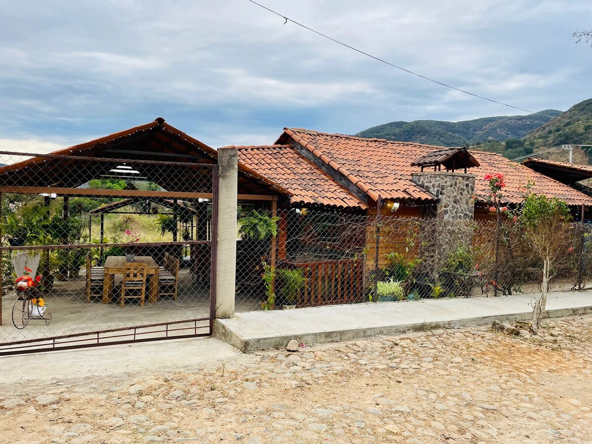 The rustic cabin is viewed from the entrance, showcasing a traditional tile roof and stone accents. Surrounding greenery is visible, along with a charming outdoor dining area featuring wooden furniture. The partially enclosed space provides a welcoming atmosphere, blending with the natural landscape.