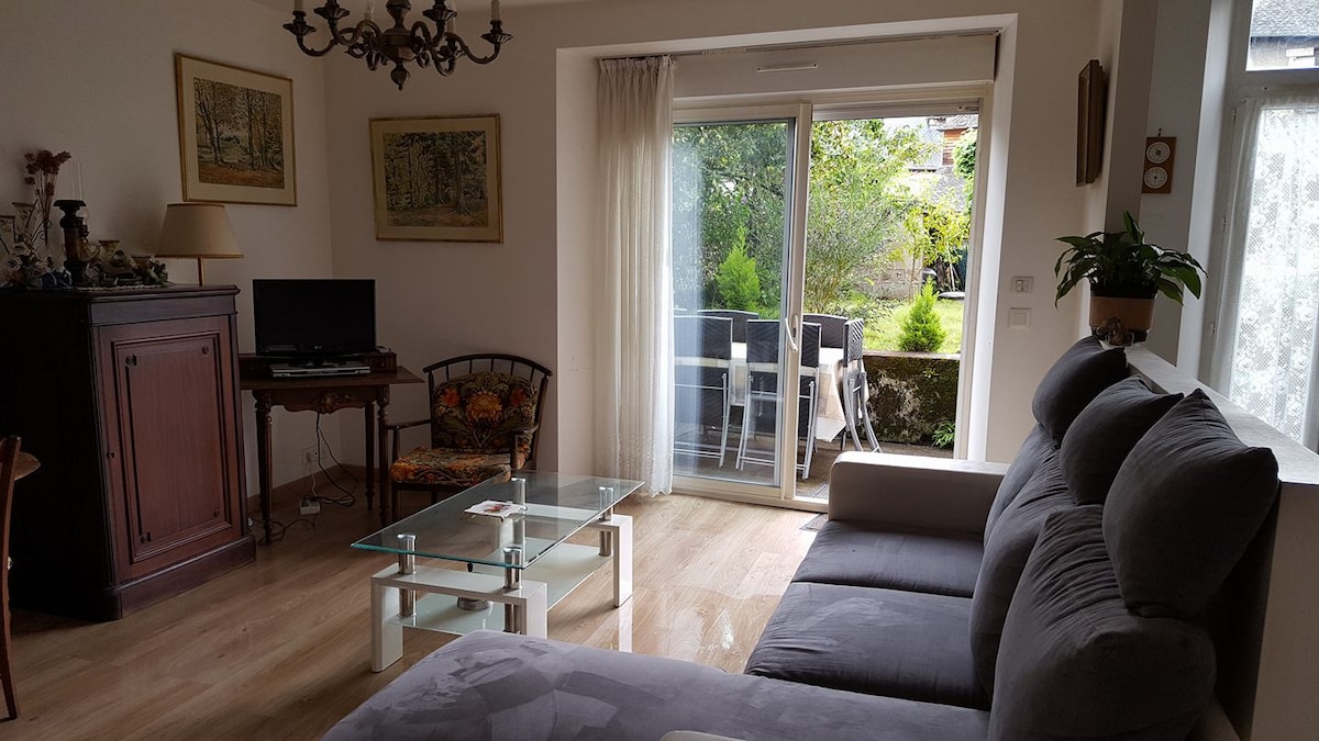 A comfortable living area is furnished with a gray sectional sofa and a glass coffee table. A television and decorative cabinet are positioned nearby. Large glass doors lead to an outdoor terrace with greenery visible beyond, enhancing the room's light and spacious feel.