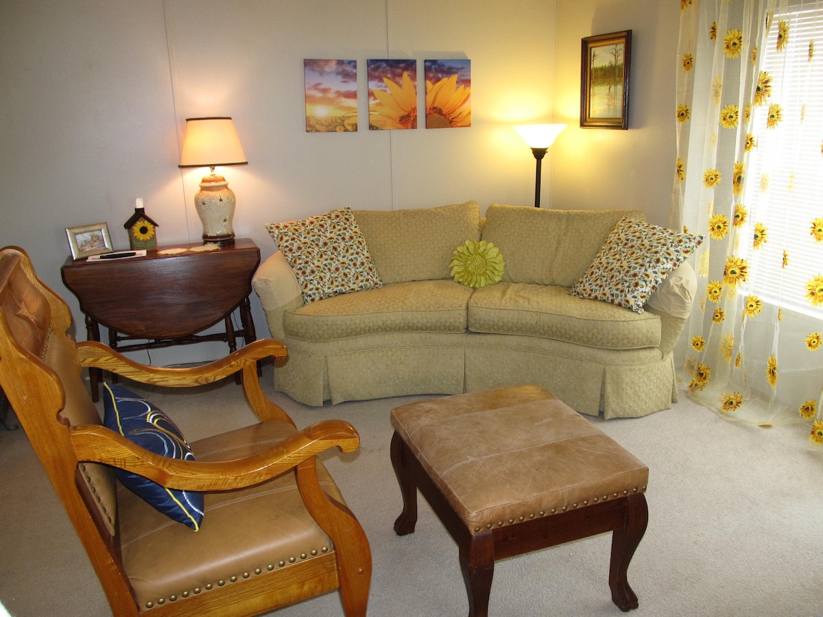 A welcoming living area features a soft yellow couch adorned with patterned throw pillows. A wooden rocking chair is positioned adjacent to a small table. Natural light filters through sunflower-patterned curtains, and soft lighting is provided by a floor lamp and a table lamp.