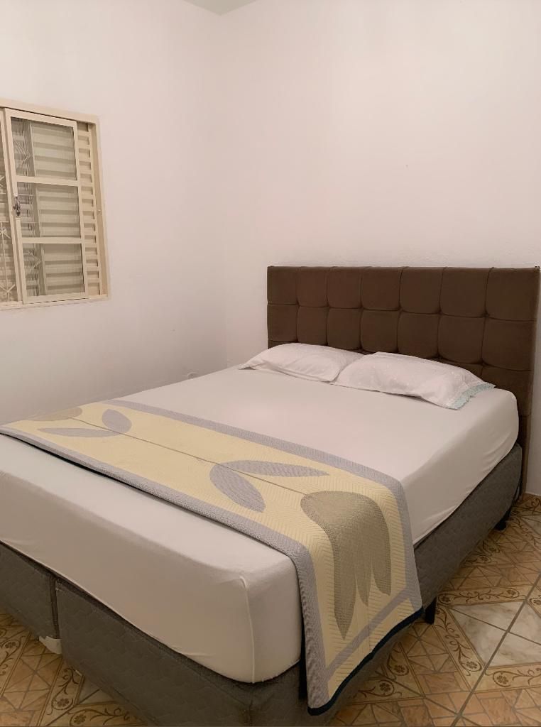 A comfortable sleeping area features a spacious bed with a soft headboard and light bedding. A light-colored blanket with a simple pattern is draped across the bed. Natural light enters through a small window with wooden shutters, illuminating the room's neutral tones.