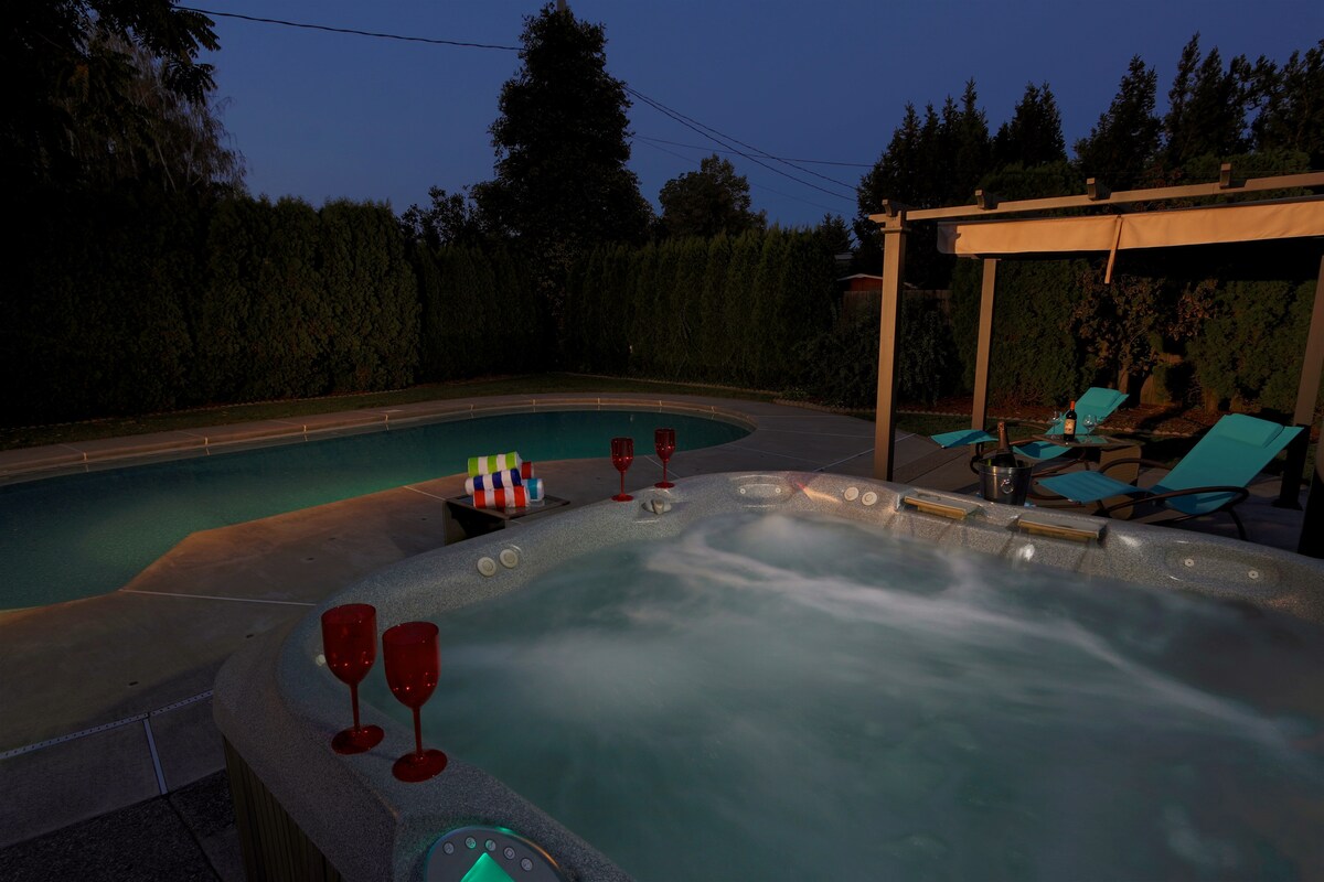 A private hot tub is positioned next to a heated pool, with bubbling water visible. Colorful chairs are arranged nearby, and a drink station featuring vibrant glasses adds a relaxing touch. Tall hedge greenery surrounds the outdoor space, enhancing privacy during evening hours.