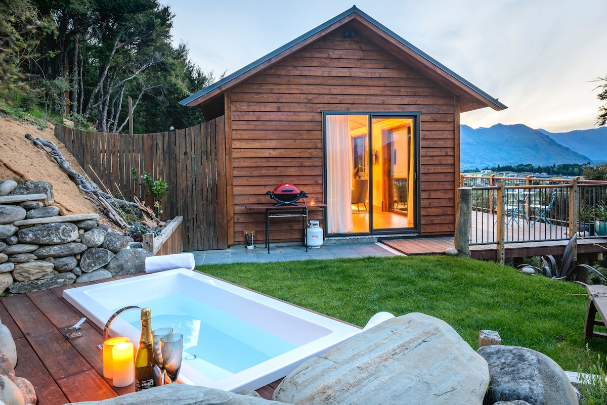 A private two-person bath is situated on a wooden deck, surrounded by large rocks and greenery. A gas grill is positioned nearby, with the chalet visible in the background, featuring wide glass doors that open to the outdoor space.
