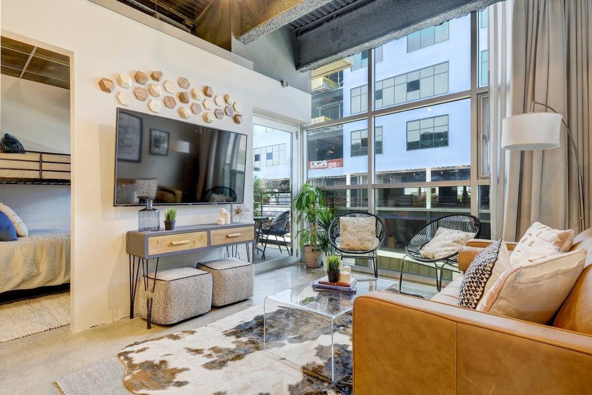 A modern living area is depicted, featuring a large mounted TV above a stylish console table. A cozy leather sofa and comfortable chairs are arranged around a glass coffee table. Natural light streams in through expansive windows, revealing views of the vibrant downtown Austin scene.