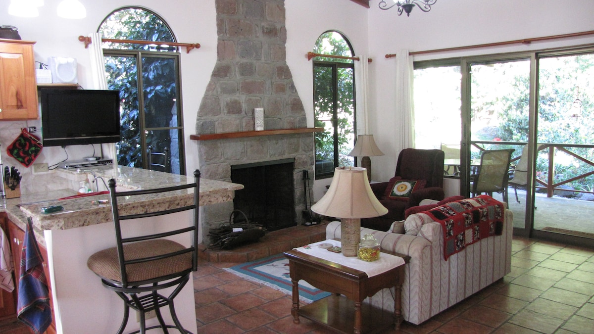An open living area is seen, featuring a stone fireplace and large windows that connect to the outdoors. A cozy seating arrangement includes a sofa and armchair, with a coffee table positioned in front. A kitchen bar with stools and a TV can also be observed.