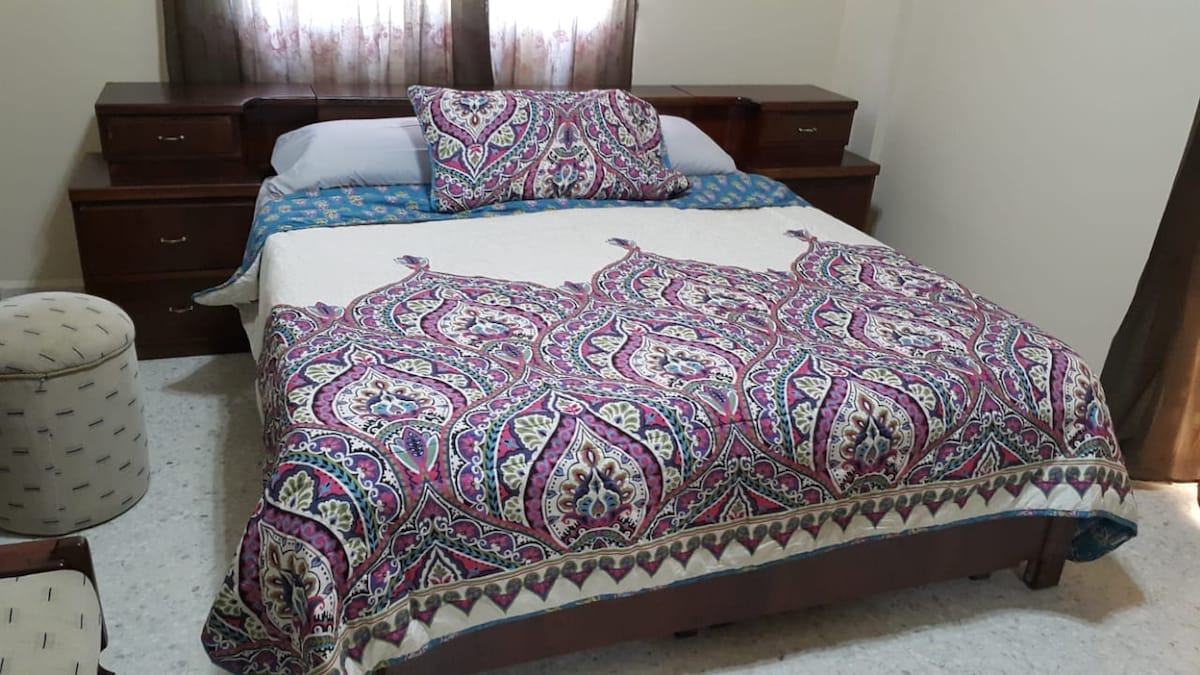 A sleeping area is furnished with a spacious bed dressed in a patterned blanket and decorative pillows. Wooden nightstands with drawers are positioned on either side, and light filters through partially closed curtains, creating a softly lit ambiance in the room.