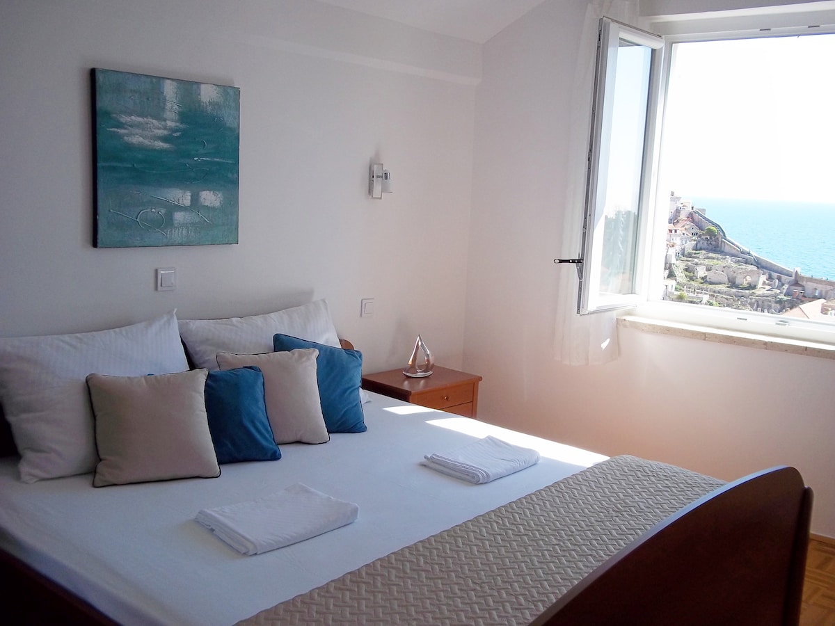 A bright bedroom is presented with a spacious bed, adorned with neutral and blue accent pillows. A large window offers a view of the sea and surrounding landscape, bringing natural light into the room. A piece of wall art complements the serene decor.