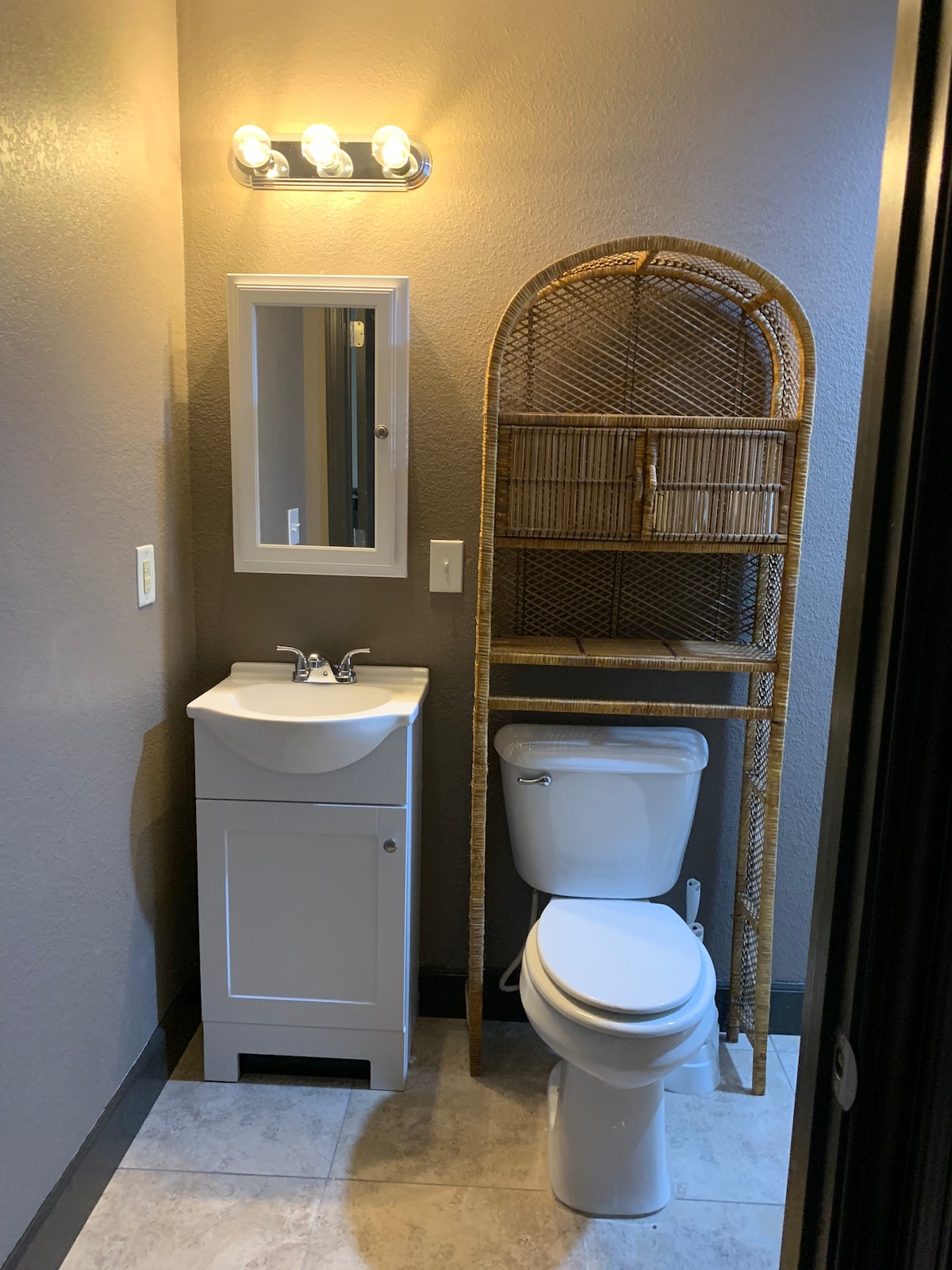 The bathroom features a compact vanity with a sink and a mirror above, accompanied by a white toilet. A woven shelf unit is positioned next to the toilet, providing additional storage space. Soft lighting is provided by a fixture mounted above the mirror.