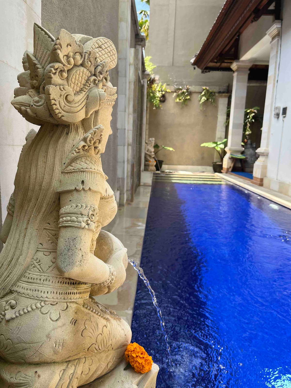 A stone statue depicting a traditional Balinese figure stands gracefully beside a sparkling private pool. The vibrant blue water reflects the surrounding greenery, creating a serene atmosphere. Sunlight filters through the area, enhancing the natural beauty of the setting.
