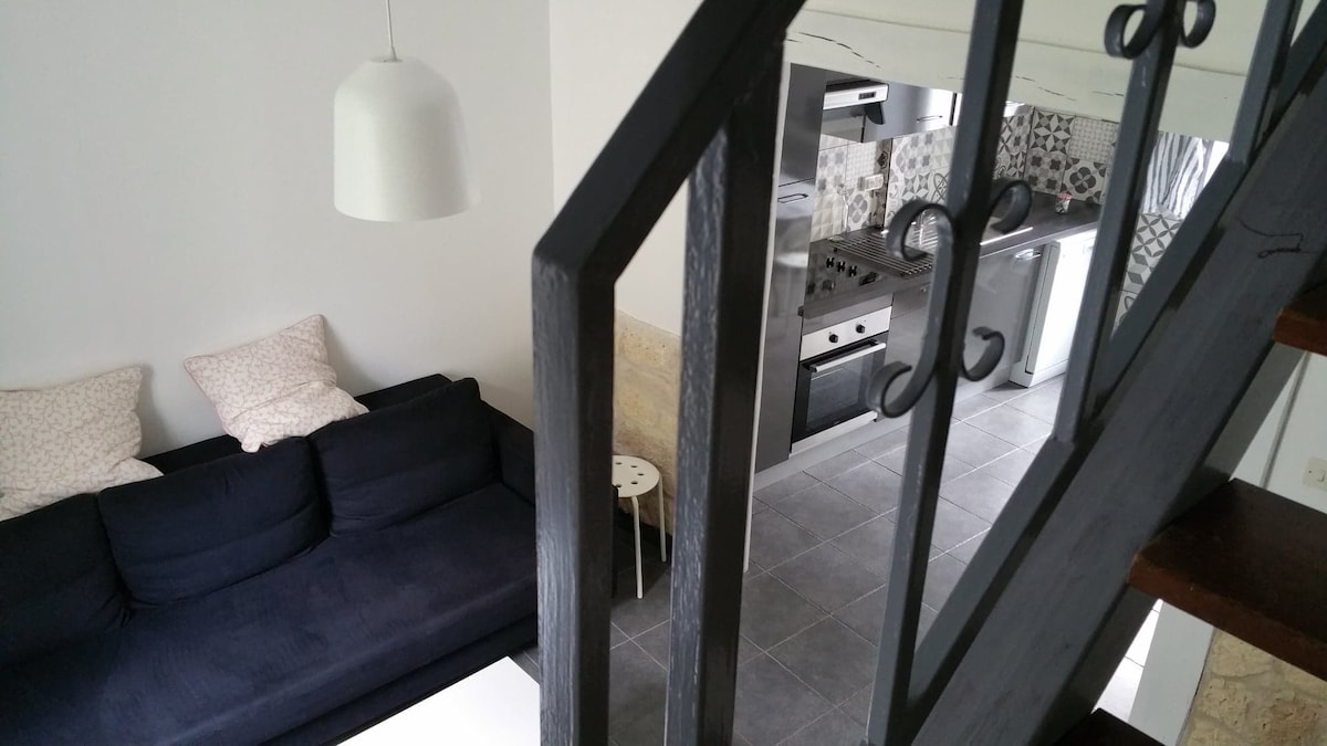 A view from above showcases a dark sofa with decorative cushions, positioned next to a small, round table. The kitchen area can be seen in the background, featuring a modern stove, and stainless steel refrigerator, with gray tile flooring throughout.