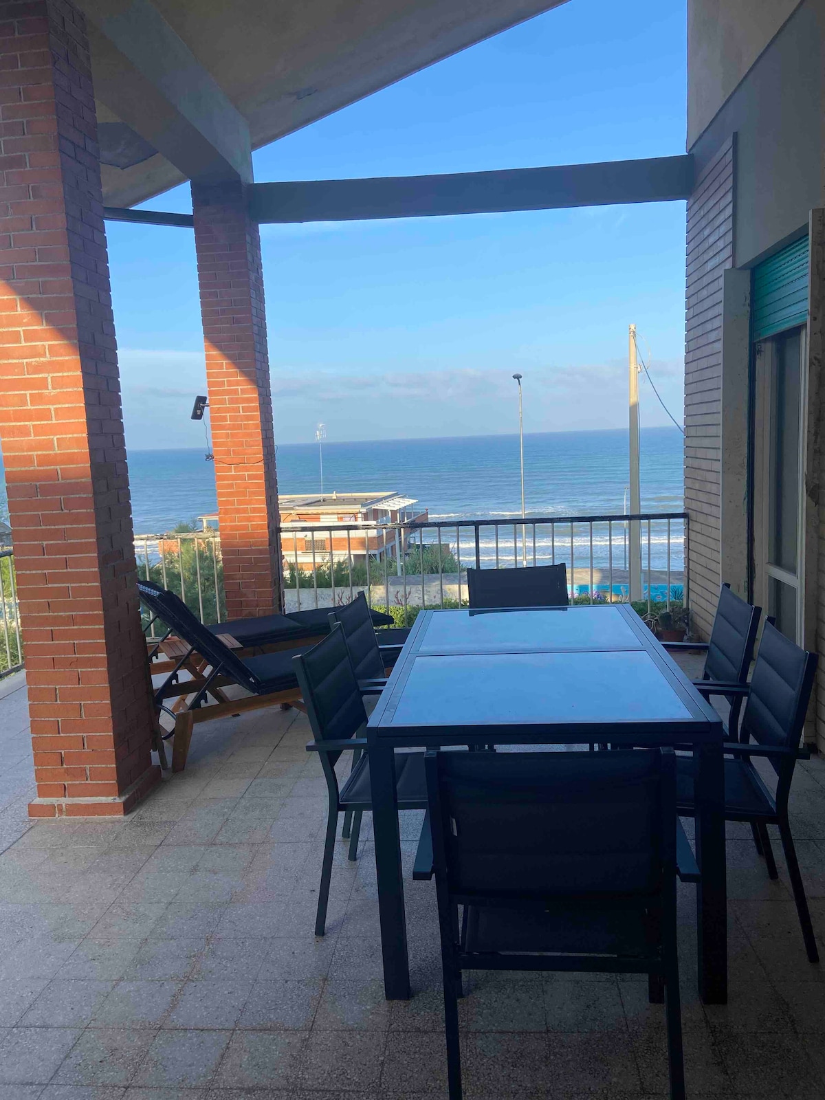 A spacious balcony offers a panoramic view of the sea, furnished with a large dining table that seats up to eight. Two lounge chairs are positioned nearby, providing a comfortable space to relax. The setting features a blend of brick pillars and tile flooring.
