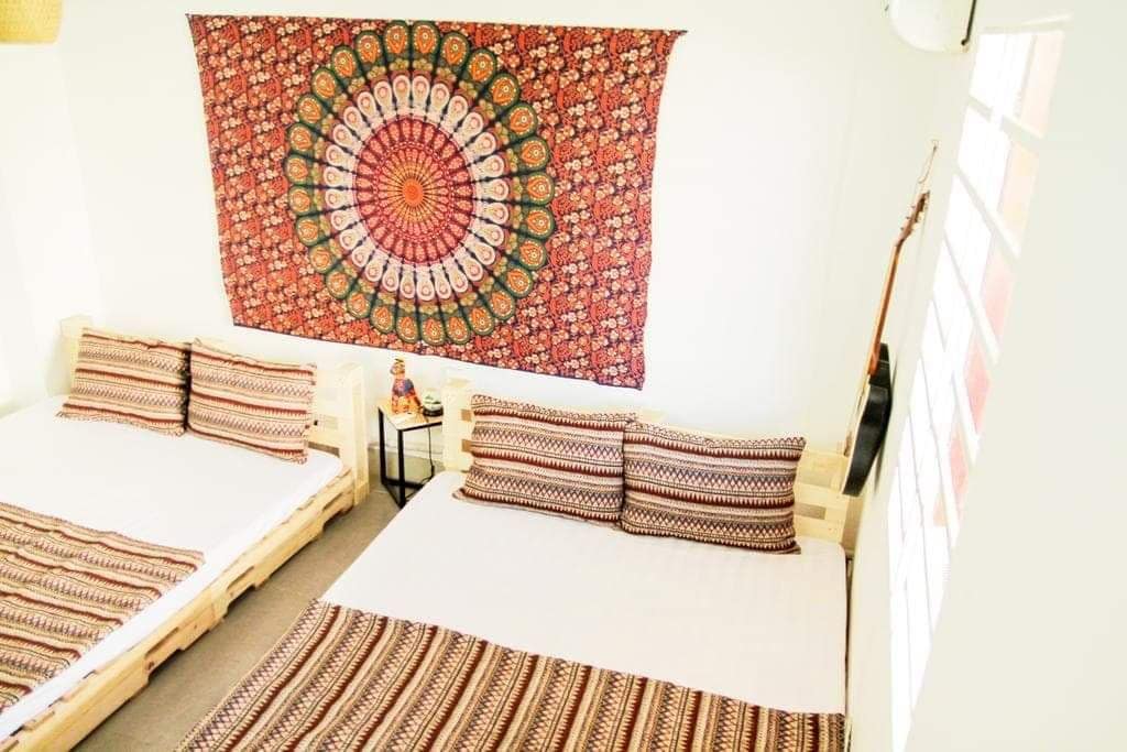 A well-lit bedroom features two spacious beds adorned with patterned blankets and decorative pillows. A colorful wall hanging with intricate designs adds visual interest, while a small table beside the beds holds decorative items, contributing to a cozy and inviting environment.