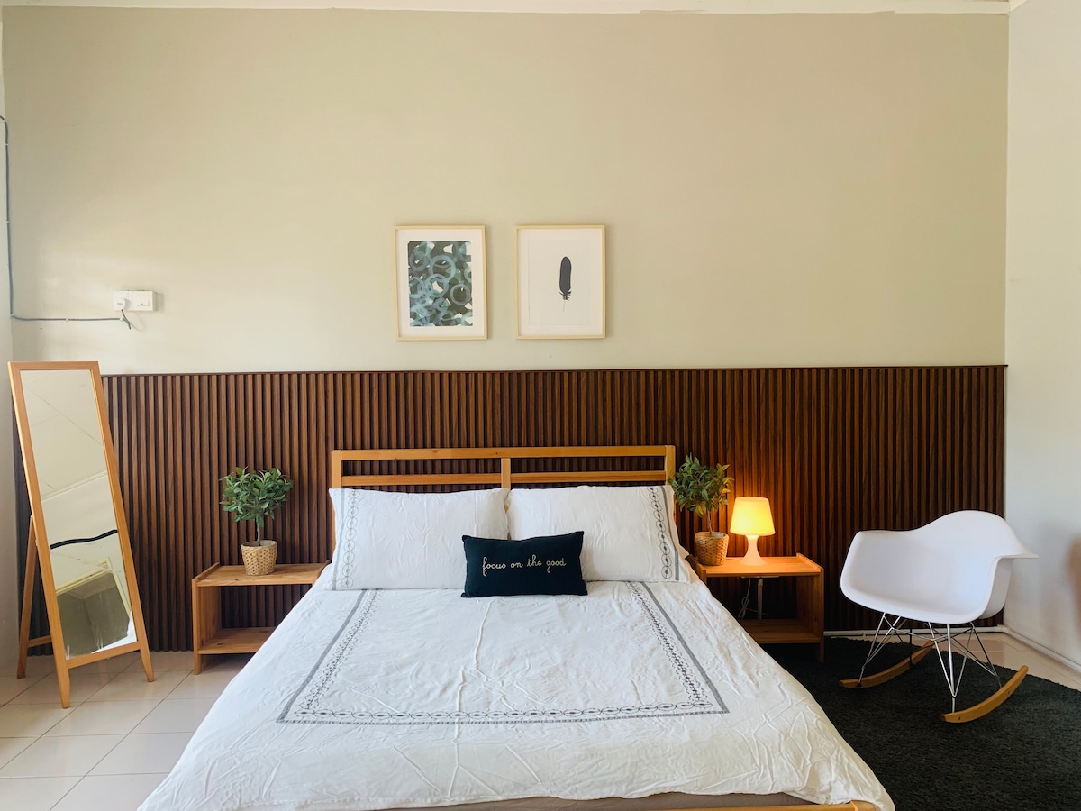 A well-lit bedroom features a spacious bed with neutral bedding. Two bedside tables with lamps are positioned alongside the bed. Green plants add a natural touch, while framed artwork decorates the wall. A large mirror and a white rocking chair provide additional decor.