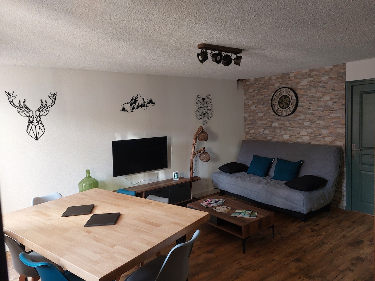 A living area is depicted, featuring a sofa bed alongside a wooden table surrounded by colorful chairs. A television is mounted on the wall, with decorative wall art including geometric designs. Natural light enhances the wood flooring, creating an inviting space.