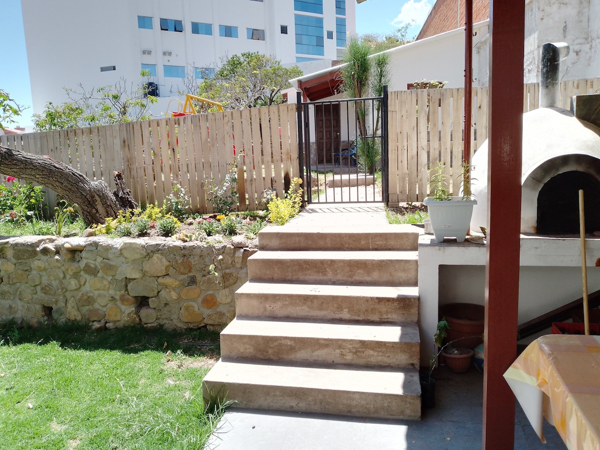 A set of stone steps leads up to a quaint garden area, featuring a wooden fence and various plants. In the background, a grill area is visible, adding to the outdoor space. Sunlight illuminates the scene, enhancing the natural greenery.