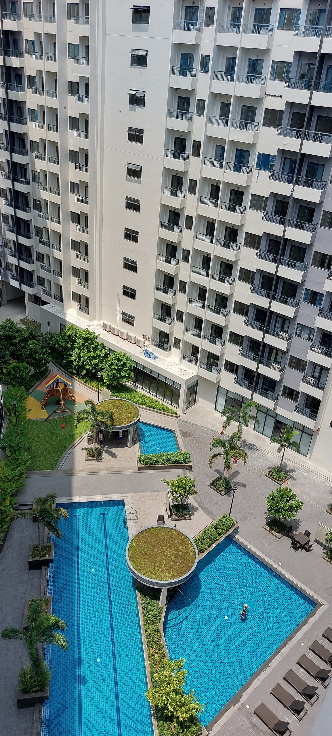 An aerial view of a landscaped area features two outdoor pools surrounded by greenery and lounge chairs. A children's playground with a small jungle gym is visible nearby, alongside manicured pathways leading to the building. The clean, modern building stands tall in the background.