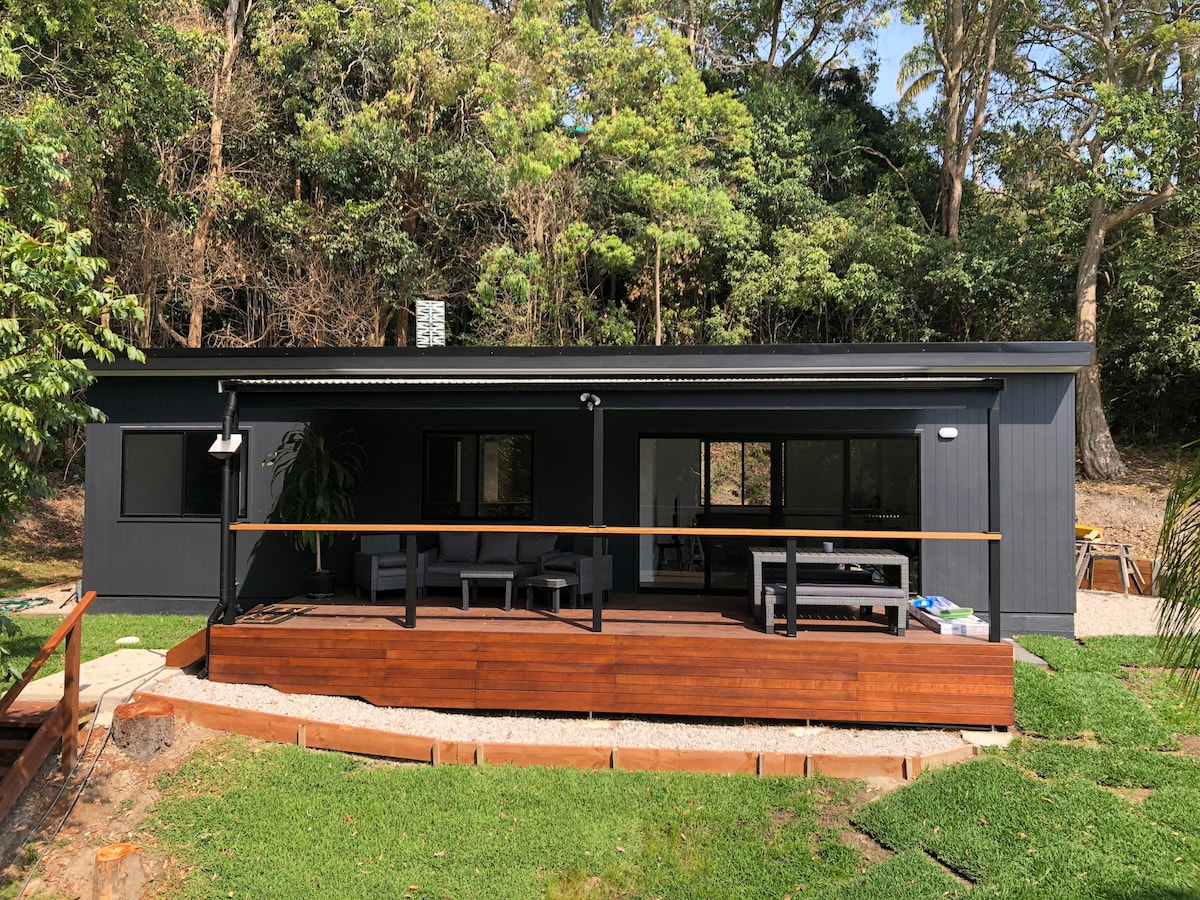 A modern guesthouse is positioned amidst lush greenery, featuring a dark exterior with large windows. A spacious deck made of wood is visible in front, offering outdoor seating options. The surrounding natural landscape enhances the secluded and tranquil environment.
