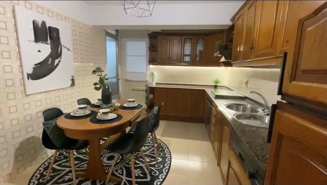 A well-appointed kitchen and dining area is depicted, featuring a round wooden table set for four with dark chairs. The cabinets display a warm wooden finish, complemented by a sleek countertop. An elegant light fixture hangs above, and modern appliances are visible in the kitchen space.