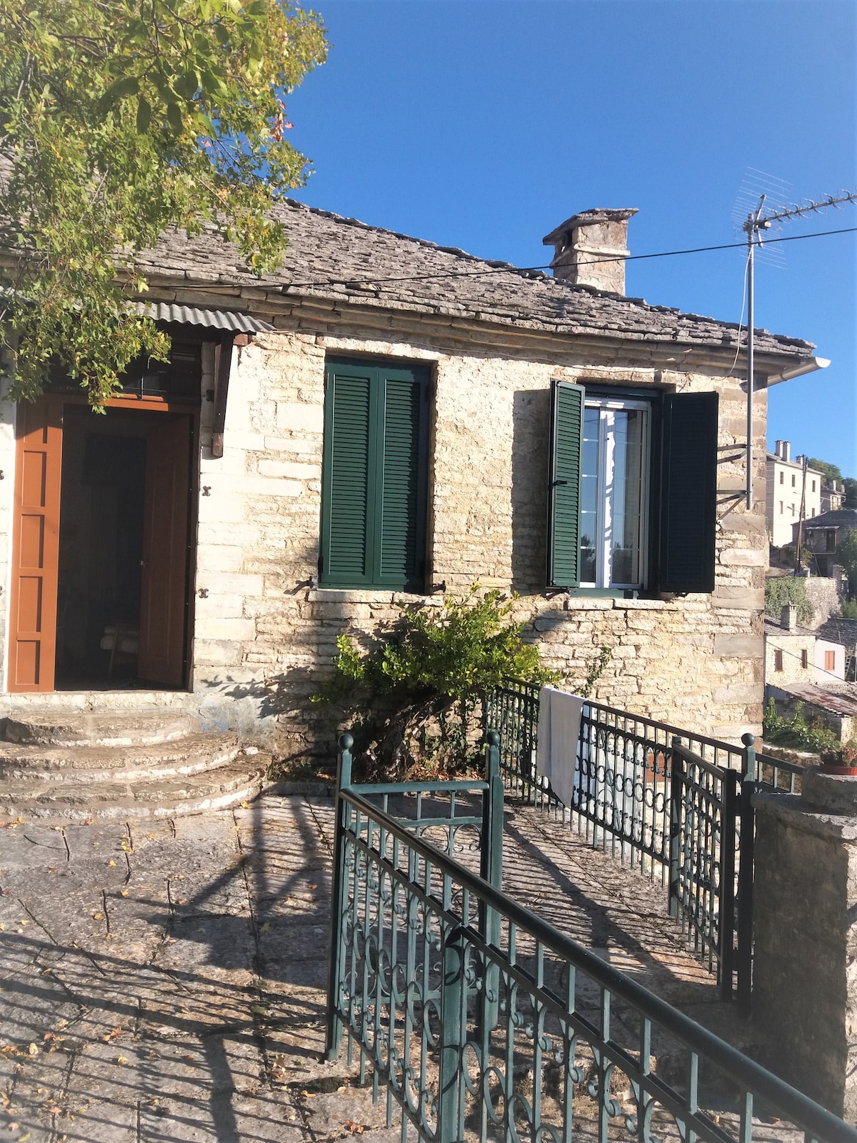 A traditional stone house features wooden shutters painted in deep green. A small set of steps leads to a welcoming entrance. To the side, a rail is partially visible, and the clear blue sky provides a bright backdrop, enhancing the home's rustic charm.