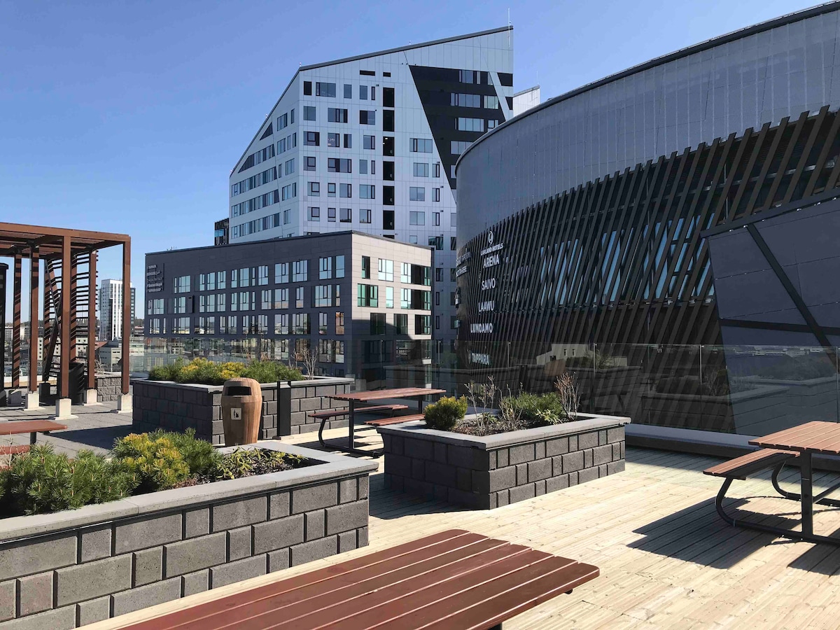 A rooftop terrace features multiple wooden picnic tables surrounded by lush greenery. Modern buildings are visible in the background, and a smooth, glass facade reflects the bright sky. The area is designed for relaxation and enjoyment, providing ample space for social gatherings.