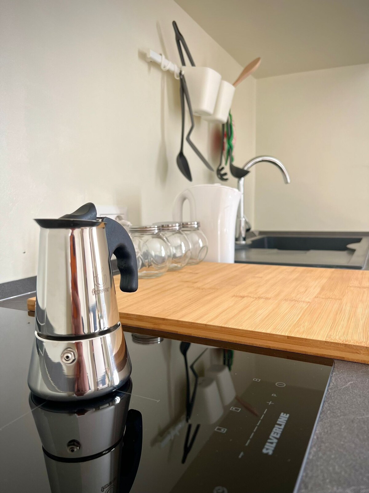 A kitchen space is presented with a modern stove and a sleek, stainless steel coffee maker prominently positioned in the foreground. A wooden cutting board rests on the countertop, with glass jars and kitchen utensils visible in the background.