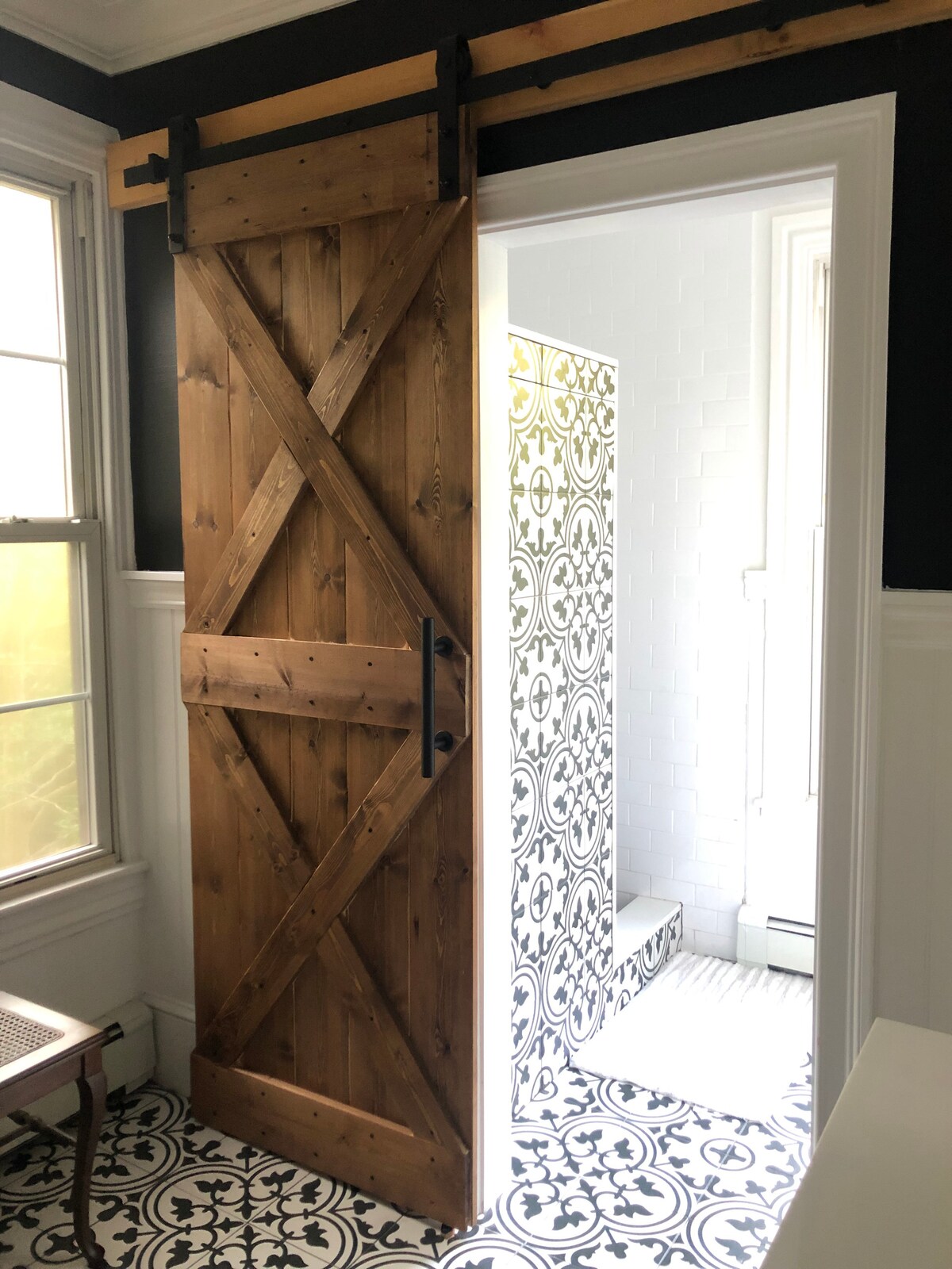 A sliding wooden barn door finished in natural tones provides access to the bathroom, framed by large windows that allow natural light. The bathroom features patterned floor tiles with black and white motifs, contrasting with sleek white tiles in the shower area.