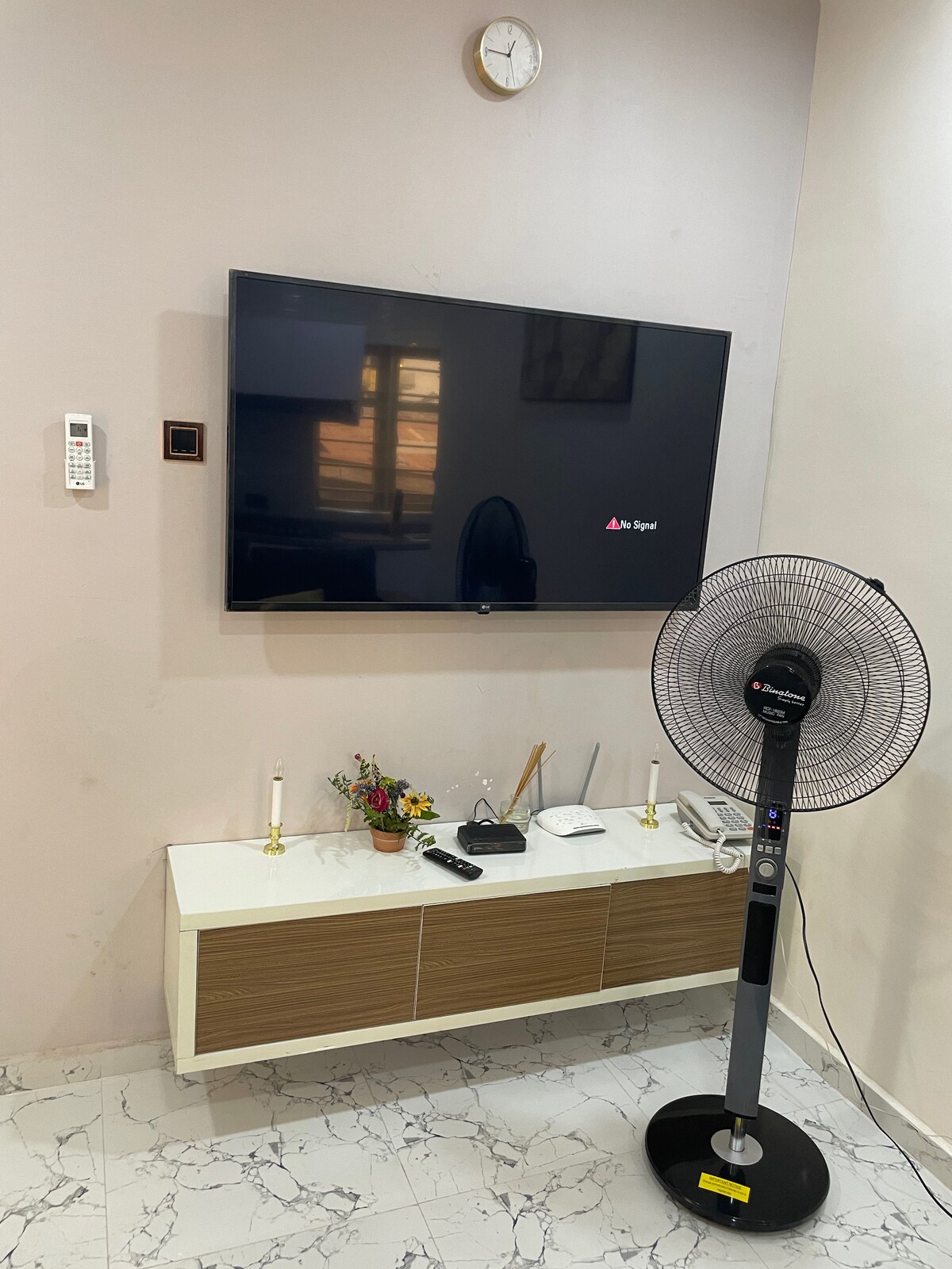 A modern entertainment space is showcased with a flat-screen television mounted on the wall above a stylish cabinet. A standing fan is positioned nearby, and decorative candles are arranged on the cabinet. Natural light filters in through a window, adding a warm ambiance.