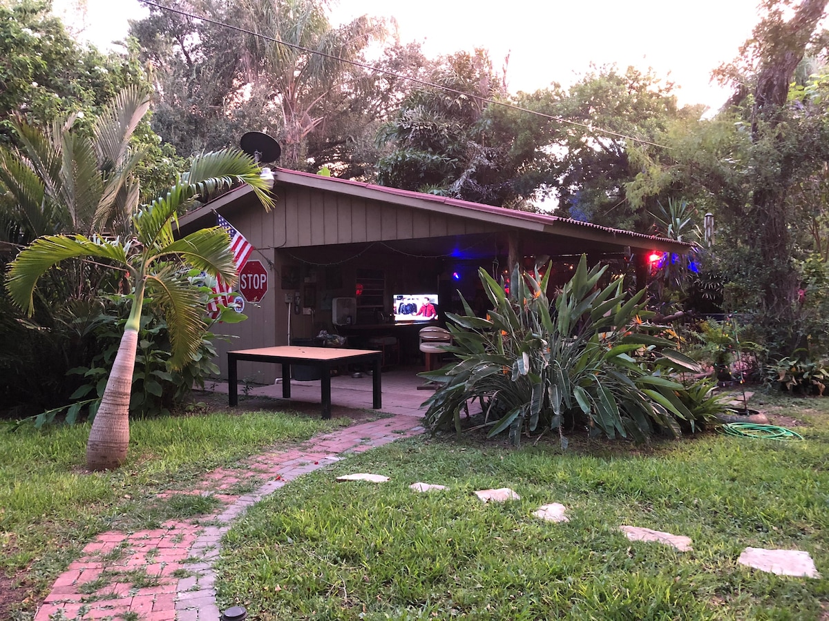The exterior of the casita is surrounded by lush greenery, featuring a combination of tropical plants and palm trees. A pathway made of stones leads to the entrance, while colorful lights add a welcoming glow in the evening. An American flag is displayed near the entrance.