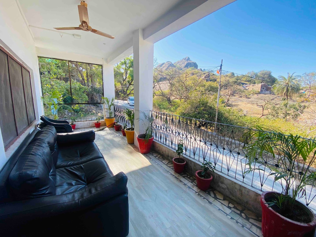 A covered terrace features black leather seating arranged for relaxation. Potted plants are positioned along the railing and floor, providing greenery. Views of the surrounding landscape are visible through the open space, showcasing trees and distant hills.