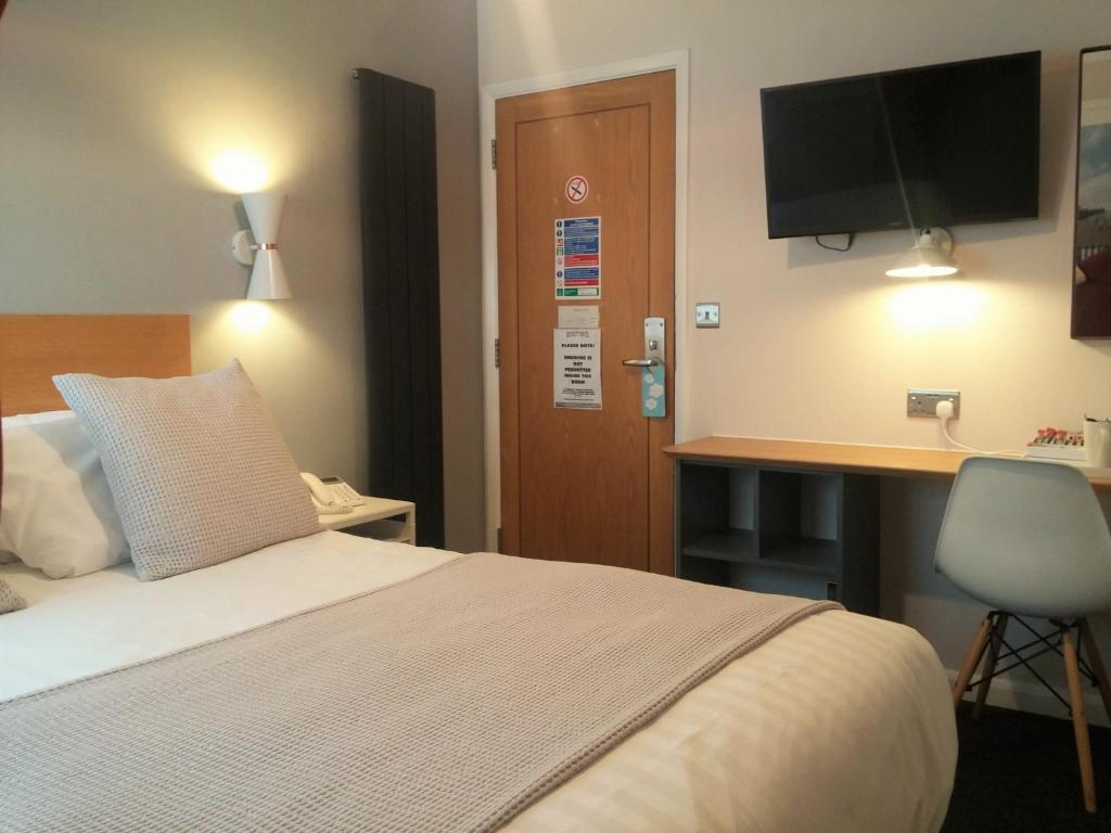 A cozy double room features a spacious bed with neutral-toned bedding and a textured throw. A small desk is positioned nearby, complemented by a modern chair. A wall-mounted television and lamps provide soft lighting, enhancing the room's functionality. The entrance door is clearly visible.