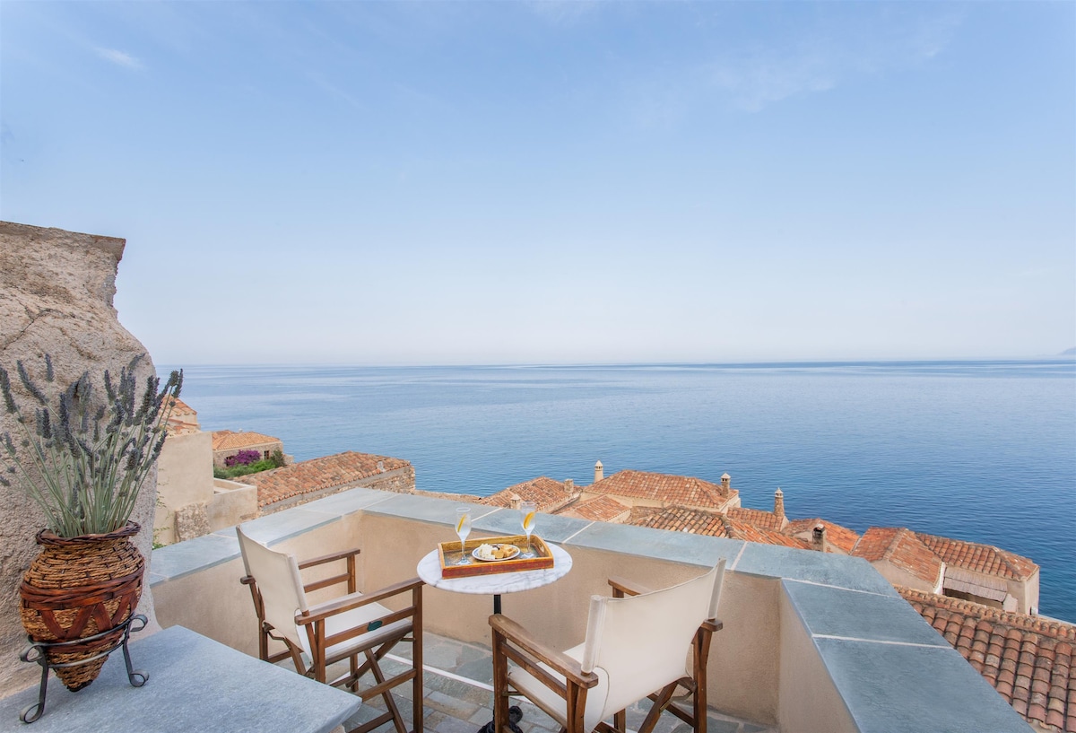 A sea view balcony is featured, showcasing two chairs and a small table. Refreshments are presented atop the table. The surrounding landscape includes a stone wall and terracotta rooftops, with calm waters extending to the horizon under a clear sky.